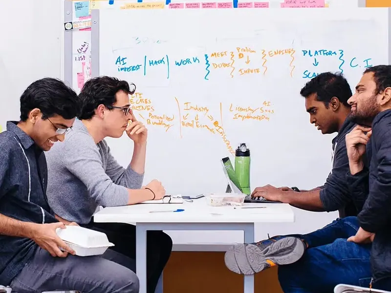 Four people working through a problem at a table in front of a whiteboard