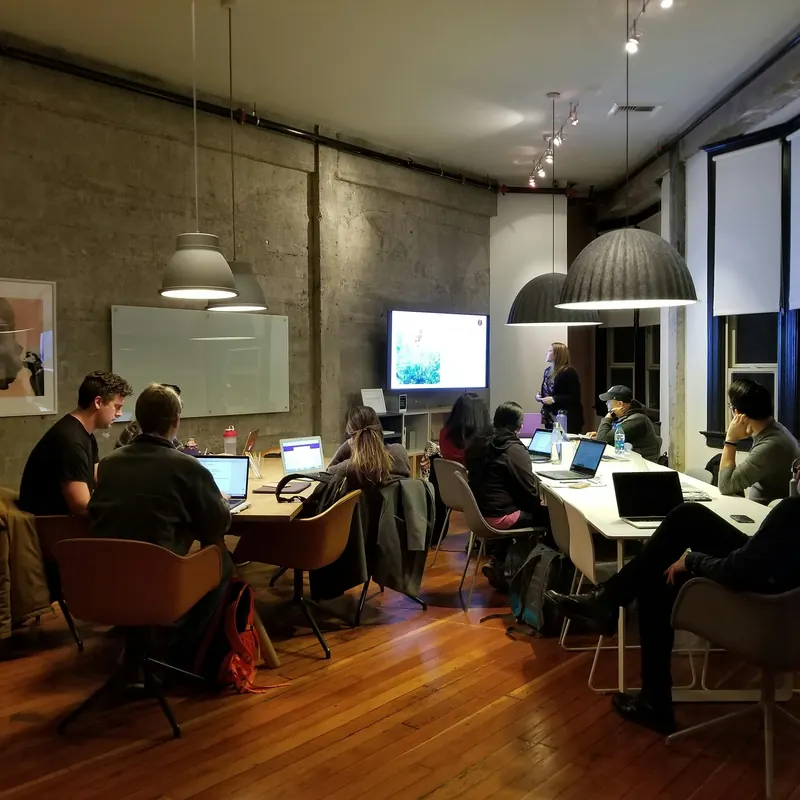 People working on laptops at long tables while a presenter speaks at a screen